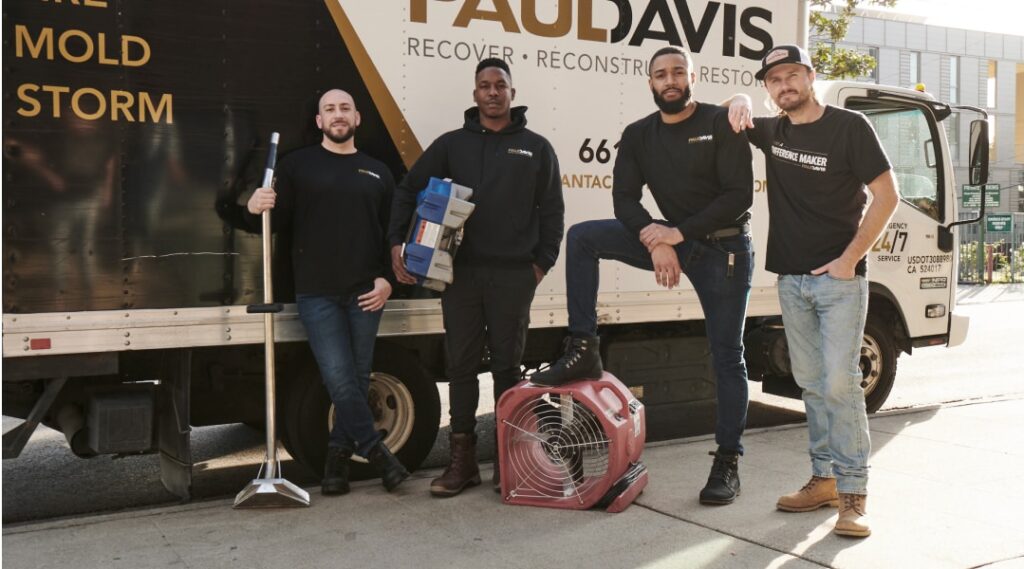 Paul Davis Restoration team members standing beside a branded service truck in Greater Myrtle Beach, SC, holding restoration equipment and posing with air mover and cleaning tools on a sunny street.